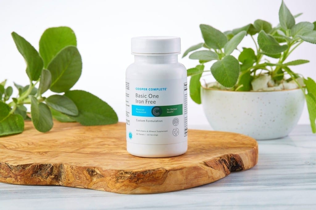 Cooper Complete Basic One Iron Free multivitamin and mineral bottle on a wood plank with cut and potted sage