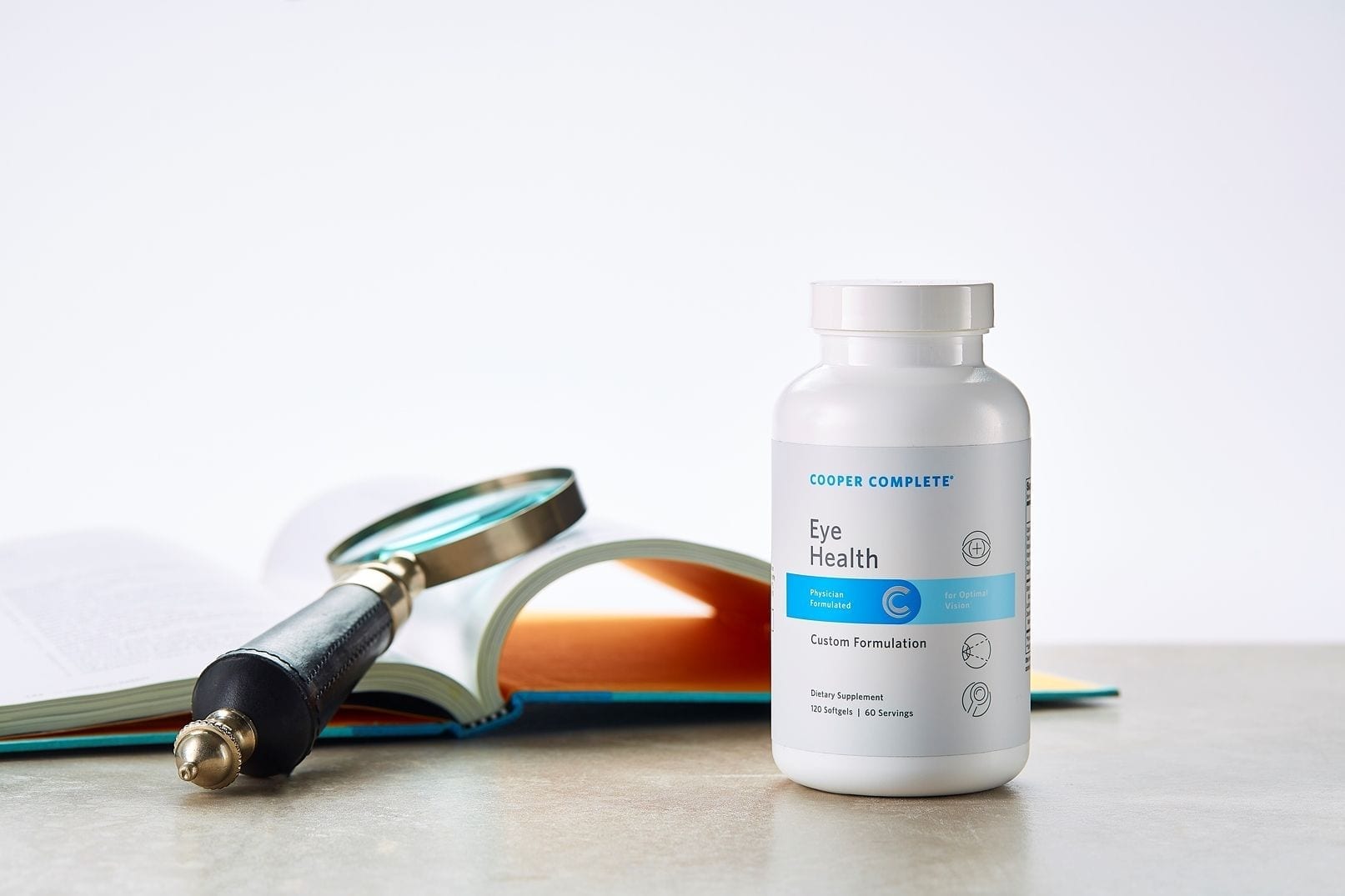 Cooper Complete Eye Health dietary supplement bottle on a tablet along side an open book with magnifying glass