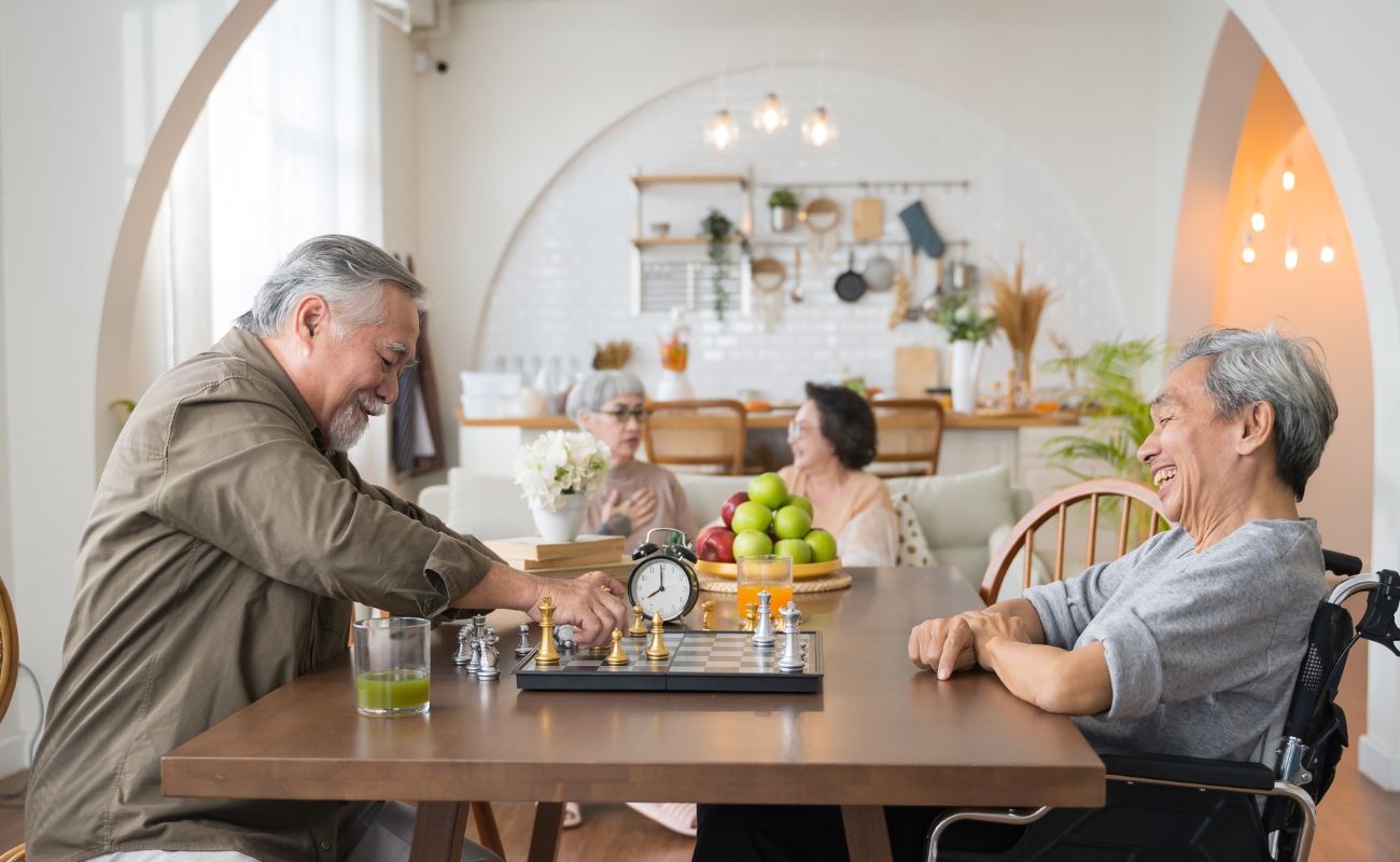 Photo of two older couples with the men playing chess at a table while the women visit on a couch nearby.
