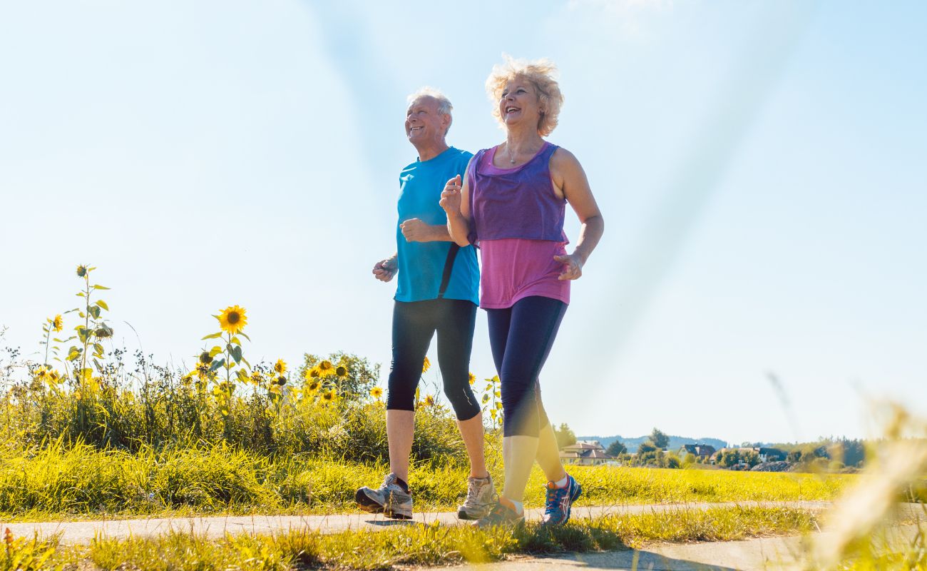 Photo of a mature man and woman in athletic wear walking on a park path past sunflowers.