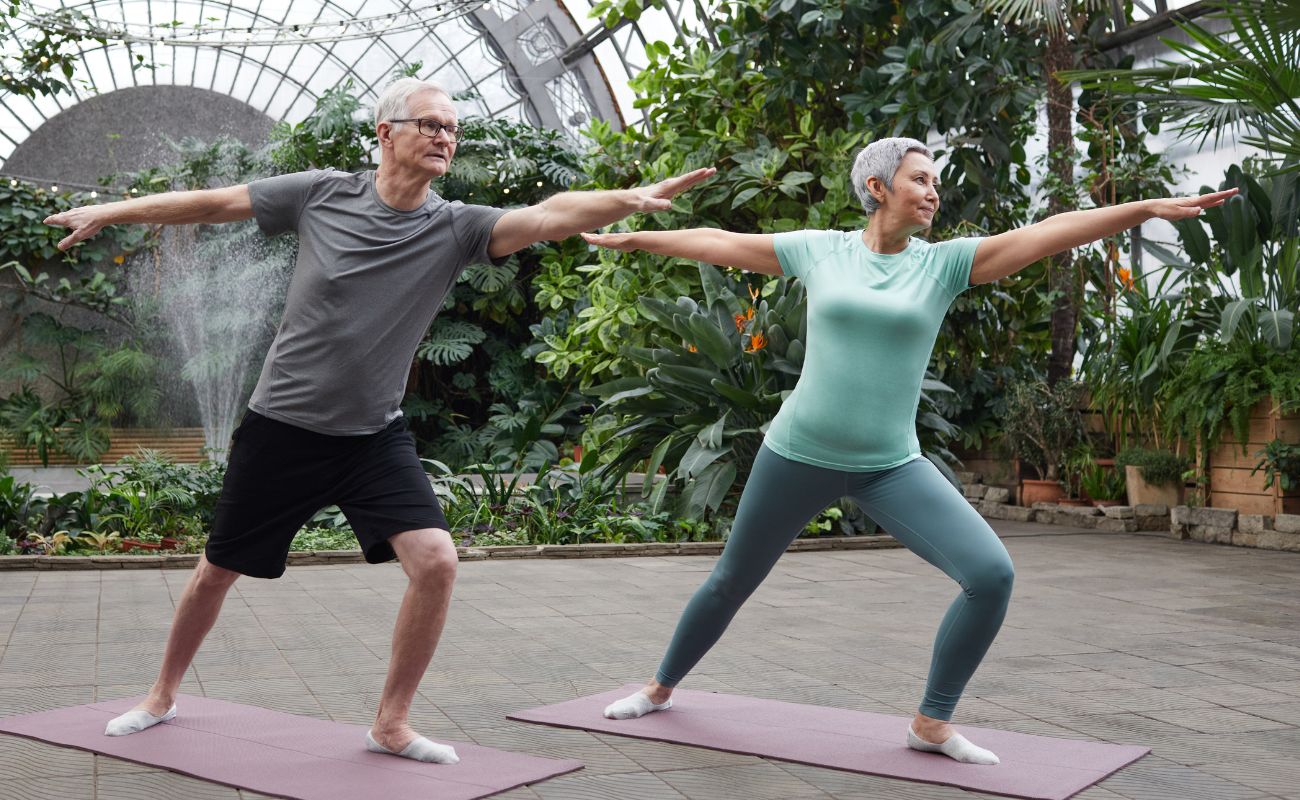 Photo of a mature couple enjoying a yoga session in a conservatory filled with plants and a water feature
