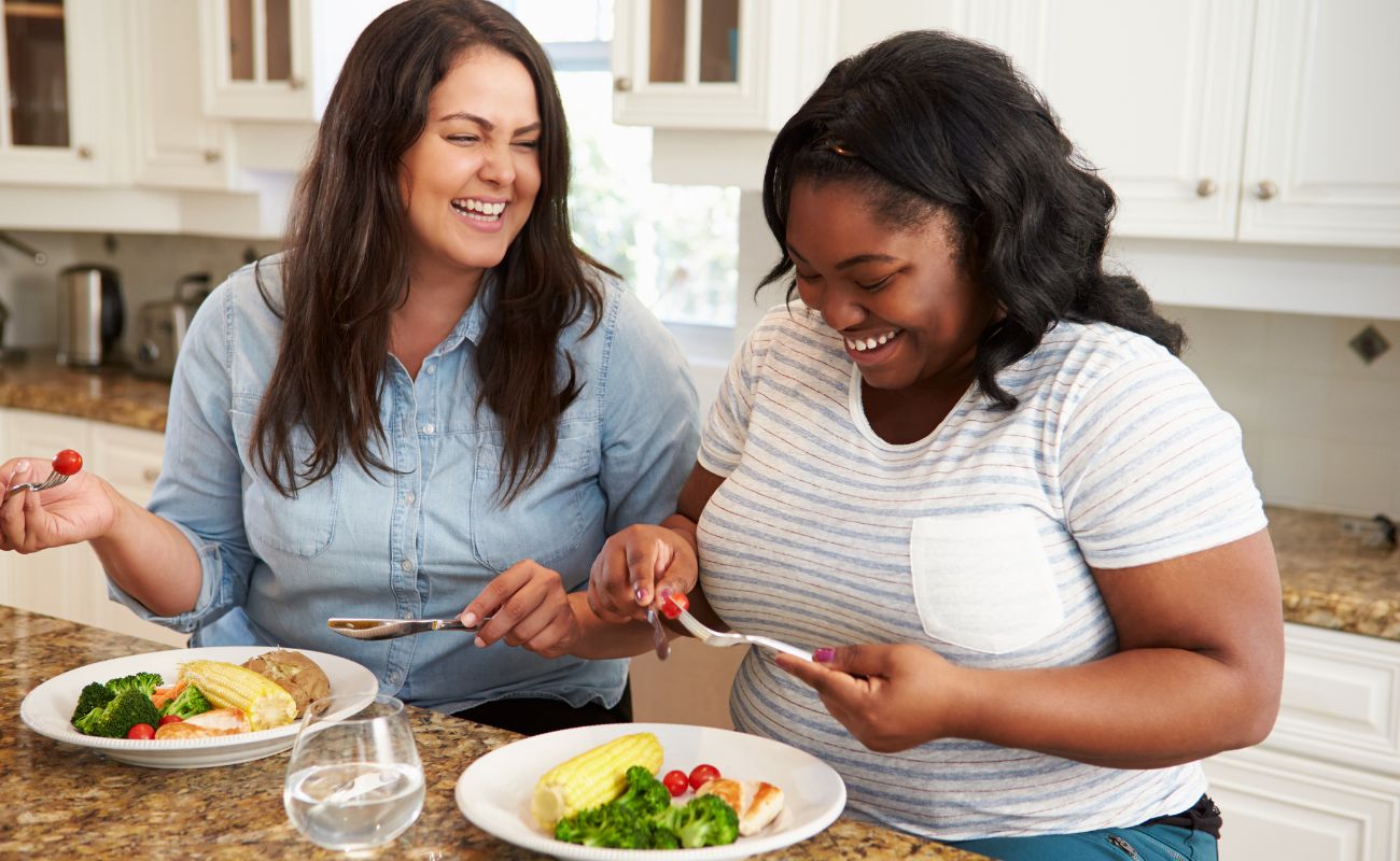 Photo of two women in the kitchen enjoying a laugh and eating a meal.
