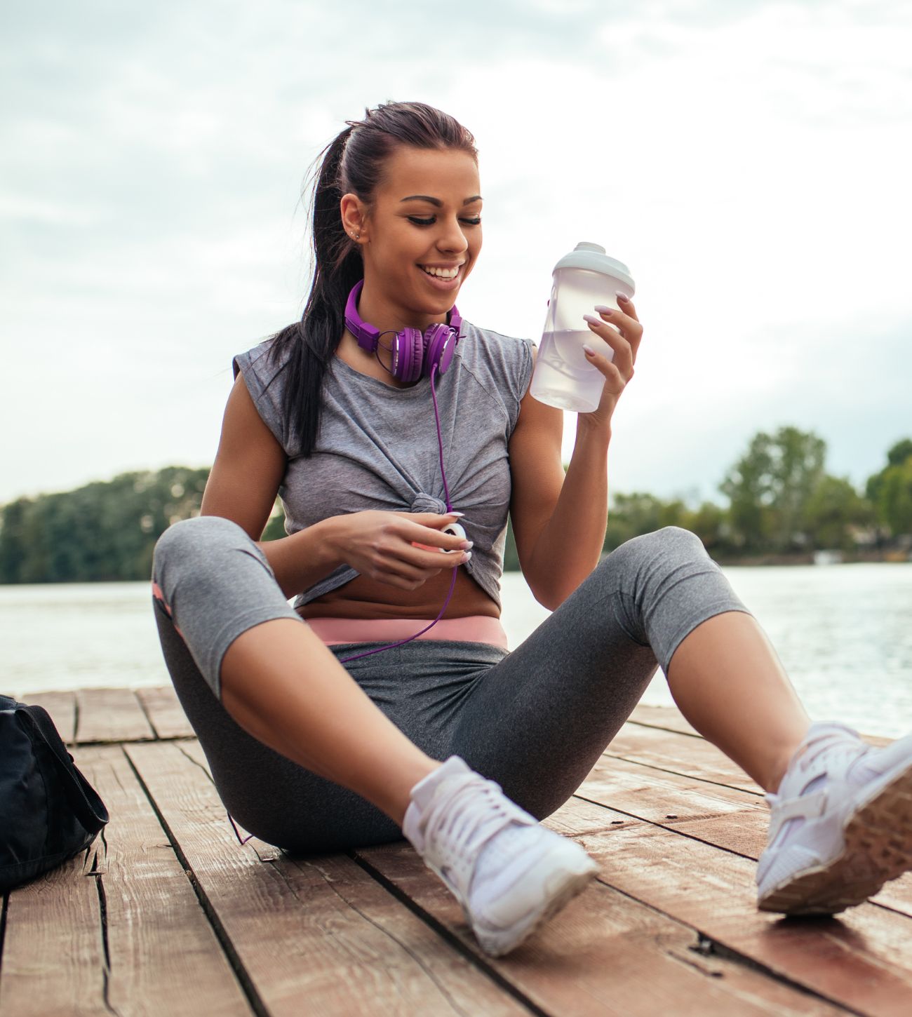 Photo of a young woman sitting on a boat dock drinking water after a workout.