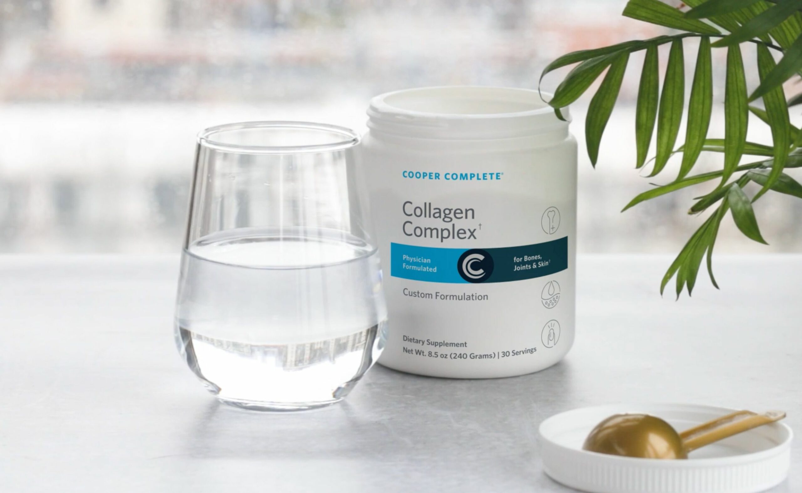 Photo of an open canister of Cooper Complete Collagen Complex beside a glass of water and a scoop.